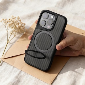 May include: A black phone case with a carbon fiber texture and a black ring holder. The case has a black frame and a circular magnetic attachment. The phone case is held by a person's hand.