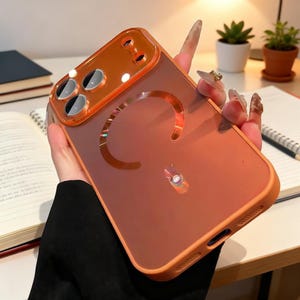 May include: Orange and clear phone case with a camera lens protector. The case has a cutout in the shape of a "C" and a circular design at the bottom. The case is held in a person's hand.
