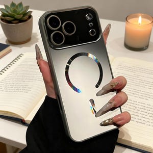 May include: A silver and clear phone case with a gradient design and a circular rainbow accent. The case has a camera cutout and is held over an open book and a lit candle.