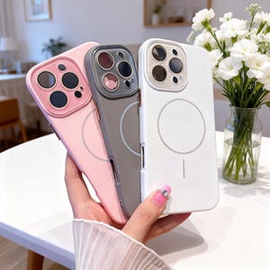 May include: Three phone cases in pink, gray, and white. Each case has a circular design on the back and a camera lens protector. The cases are held in a hand, showcasing their design and color variations.