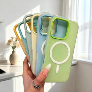 May include: A collection of colorful phone cases in shades of yellow, teal, blue, and green. Each case has a white circular design and a matching colored border around the camera. The cases are held in a hand.