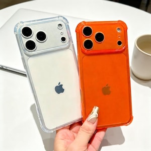 May include: Two phone cases, one clear with a white back and the other orange. Both cases have a protective bumper and a cutout for the camera lenses. The Apple logo is visible on the back of each case.