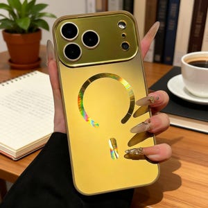 May include: A gold-colored phone case with a reflective, iridescent design. The case has a camera cutout and a circular design on the back. The phone case is held in a hand.