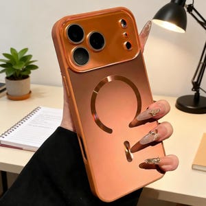 May include: A copper-colored phone case with a glossy finish. The case features a raised camera section and a circular design on the back. The case is designed to protect the phone and has a built-in ring holder.