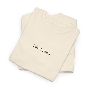 May include: Two cream-colored t-shirts are stacked on top of each other. The text "i do brows" is printed in black on the front of the top shirt. The shirts are made of a soft, textured material.