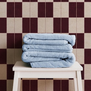 May include: A stack of three folded light blue towels resting on a white stool. The background features a wall with a repeating checkered pattern of dark maroon and beige rectangular tiles arranged in a vertical grid layout.