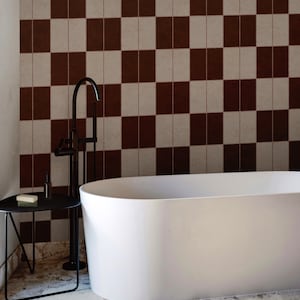 May include: A white freestanding oval bathtub positioned in front of a wall with a brown and beige checkered tile pattern. A black metal side table holds a small bottle and a bar of soap, next to a tall black floor-mounted bathtub faucet.