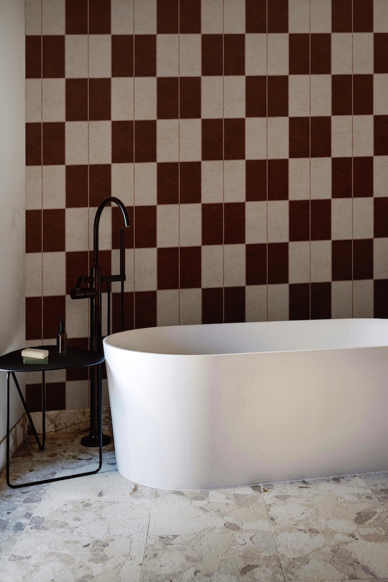 May include: A white freestanding oval bathtub positioned in front of a wall with a brown and beige checkered tile pattern. A black metal side table holds a small bottle and a bar of soap, next to a tall black floor-mounted bathtub faucet.