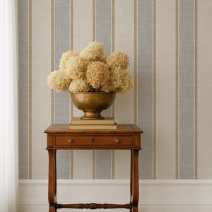 May include: A wooden side table with a drawer and a brass bowl filled with dried flowers. The table is set against a striped wallpaper with vertical stripes in beige, blue, and brown. A white curtain is visible on the left.