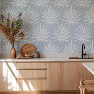 Puede incluir: Una cocina moderna con gabinetes de madera clara y un papel tapiz azul claro con un patrón de hojas de palma blancas. Un grifo negro y una tabla de cortar de madera están en la encimera. Una alfombra tejida y un taburete de madera completan la escena.