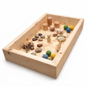 Puede incluir: Una bandeja sensorial de madera llena de elementos naturales. Incluye bloques de madera, una bola, bolas de fieltro, piedras y una lupa. La bandeja es de madera clara y está sobre una superficie blanca. También se incluye una colección de vainas secas.
