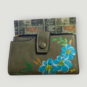 May include: Olive green wallet with a snap closure, featuring hand-painted blue flowers with yellow centers and green leaves. The wallet has a zippered compartment and a decorative floral design.
