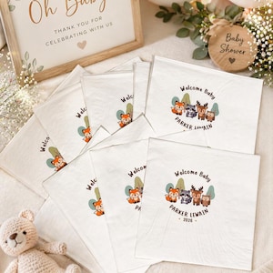 May include: A collection of white baby shower napkins with a woodland animal design and the text "Welcome Baby PARKER LEWAIN 2026". A wooden sign reads "Oh Baby!" and a wooden disc says "Baby Shower". A stuffed bear is also present.