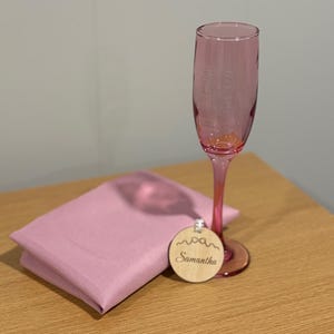 Pode incluir: Uma taça de champanhe rosa, um guardanapo rosa dobrado e uma etiqueta de madeira com o nome "Samantha" são dispostos sobre uma superfície de madeira. A taça e o guardanapo são de um tom rosa correspondente.