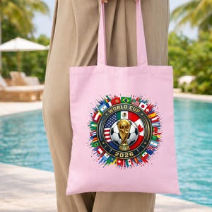 May include: A light pink tote bag featuring a colorful design for the 2026 World Cup. The design includes a soccer ball, the World Cup trophy, and flags from various countries. The bag has matching pink handles.