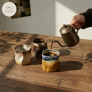 Könnte beinhalten: Drei Keramik-Kaffeetassen mit rustikalem Design. Eine Tasse wird mit Kaffee aus einem Metall-Pour-Over-Kessel gefüllt. Die Tassen haben eine strukturierte, organische Form und eine Mischung aus braunen, beigen und blau-gelben Farben. Das HomeDetails-Logo befindet sich oben links.