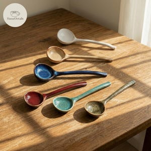 May include: A collection of six ceramic spoons in various colors, including white, tan, blue, red, teal, and olive green. The spoons are arranged on a wooden surface, showcasing their unique shapes and finishes. The spoons are ideal for serving or eating.