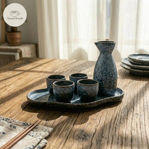 Pode incluir: Um conjunto de saquê japonês com acabamento azul escuro salpicado. O conjunto inclui uma garrafa, quatro xícaras pequenas e uma bandeja de servir. Os itens estão dispostos em uma mesa de madeira, com uma toalha de mesa e pauzinhos em primeiro plano.