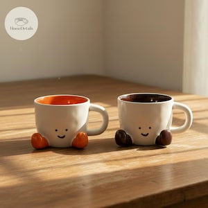 May include: Two white ceramic mugs with a smiling face design and small feet. One mug has an orange interior, the other has a brown interior. The mugs are on a wooden surface.