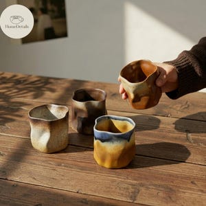 Pode incluir: Quatro canecas de cerâmica com formas irregulares e tons terrosos são exibidas em uma superfície de madeira. As canecas variam em cor, com tons de marrom, bege e azul. Uma caneca está sendo segurada por uma pessoa. O logotipo HomeDetails é visível.