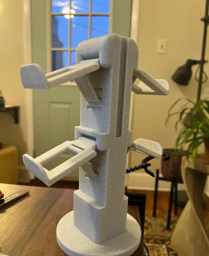 May include: A light grey, 3D-printed stand with multiple angled arms, designed to hold items. The stand has a circular base and a tall, rectangular central structure. The arms extend outwards at various levels, creating a multi-tiered design.