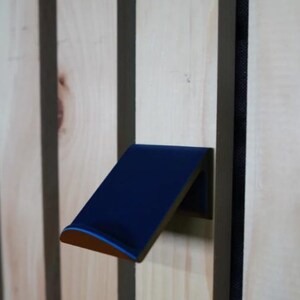 Puede incluir: Un gancho de pared azul marino en &aacute;ngulo con un borde redondeado, montado en un panel de madera clara con rayas verticales marr&oacute;n oscuro. El gancho tiene un fino acento azul claro en el borde.
