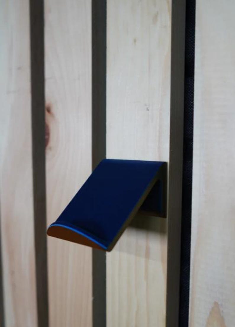 Puede incluir: Un gancho de pared azul marino en &aacute;ngulo con un borde redondeado, montado en un panel de madera clara con rayas verticales marr&oacute;n oscuro. El gancho tiene un fino acento azul claro en el borde.