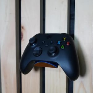 Puede incluir: Un mando Xbox negro con botones de colores, montado en una pared de madera. El mando tiene un dise&ntilde;o elegante con una superficie texturizada y est&aacute; sujeto por un soporte con un acento azul y naranja.