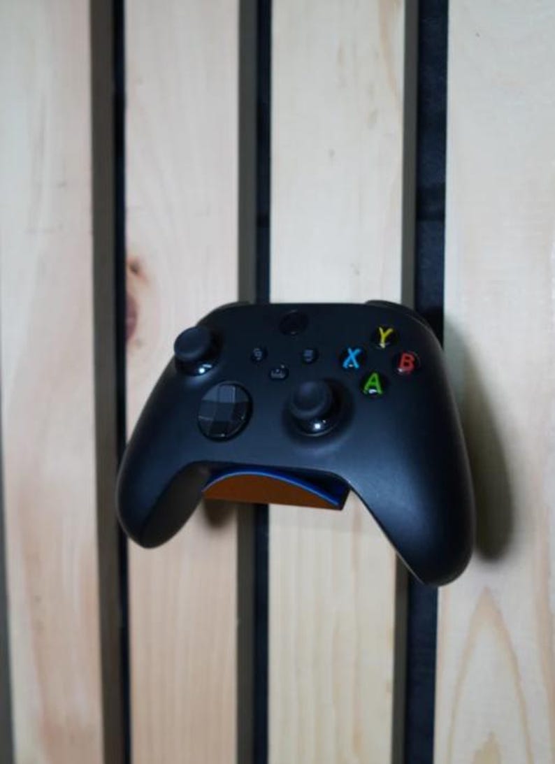 Puede incluir: Un mando Xbox negro con botones de colores, montado en una pared de madera. El mando tiene un dise&ntilde;o elegante con una superficie texturizada y est&aacute; sujeto por un soporte con un acento azul y naranja.
