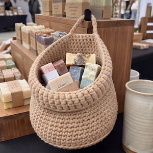 May include: A beige crocheted hanging basket filled with various rectangular bars of soap. The basket features a thick, textured knit pattern and a sturdy handle. Several additional bars of soap wrapped in paper bands are arranged on wooden shelves nearby.