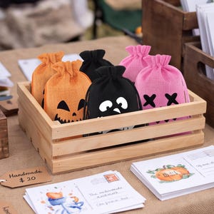 May include: A collection of small drawstring pouches made of textured fabric in orange, black, and pink. The orange pouches feature a jack-o'-lantern face, the black pouches display a ghost-like character, and the pink pouches show an 'X' eye design.