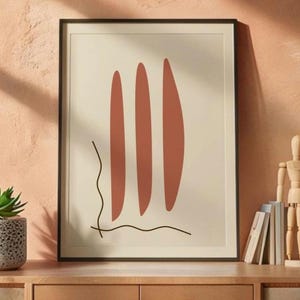 May include: A framed wall art print featuring three vertical terracotta-colored elongated oval shapes centered on a cream background, with a thin, dark brown wavy line extending from the bottom left corner across the lower section.