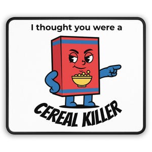 May include: A white mouse pad with a black border featuring a cartoon cereal box character. The red box has blue stripes, arms, and legs, and a bowl of cereal. Text reads "I thought you were a CEREAL KILLER."