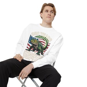 May include: White sweatshirt featuring a green lizard with yellow eyes, set against an American flag design. The text reads "250 YEARS OF REGROW WHAT'S LOST". The sweatshirt is a casual style.