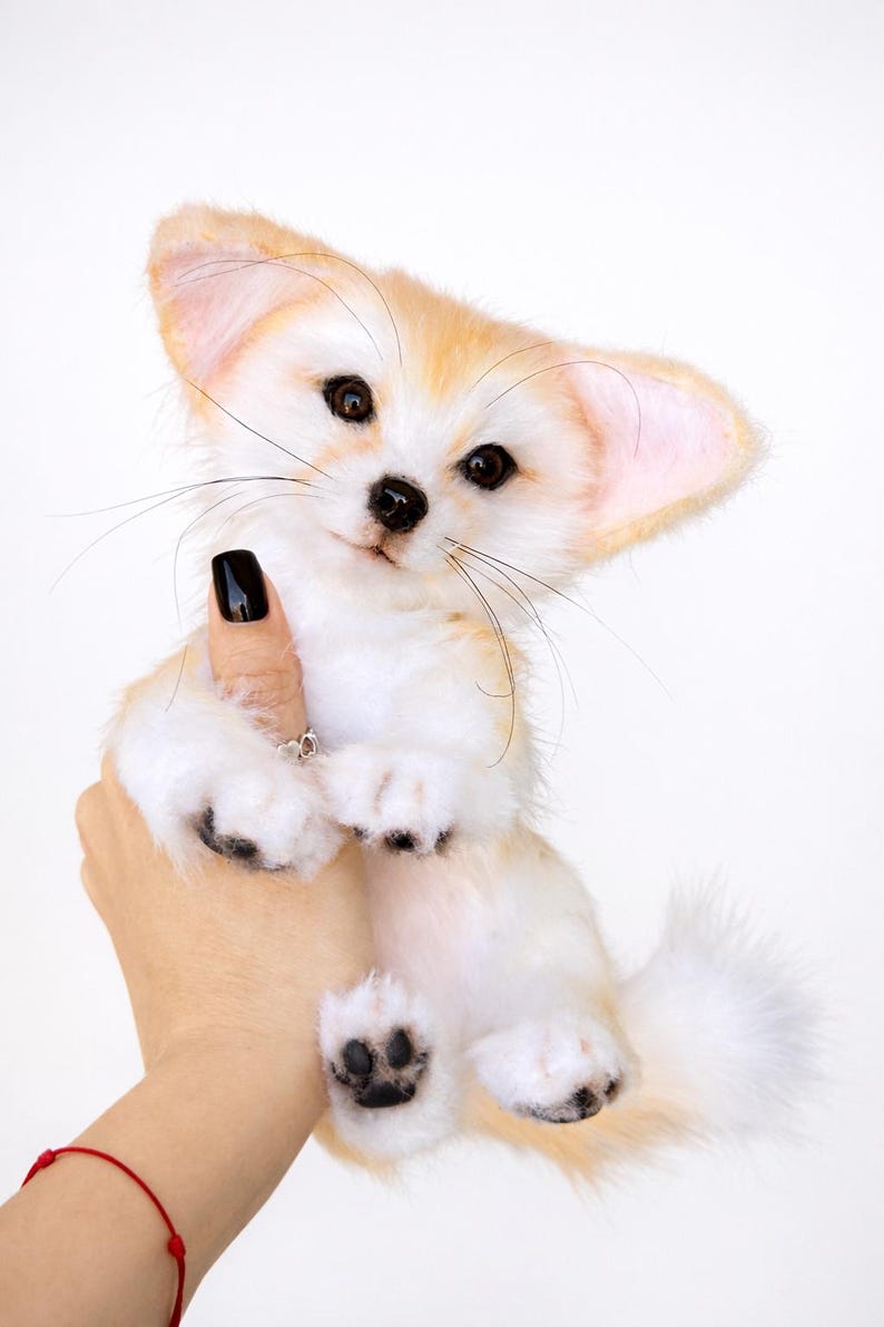May include: A plush Fennec fox toy with large ears, a white body, and tan accents. The toy has black paws and a fluffy tail. The fox is being held up against a white background.