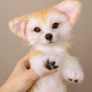 May include: A plush Fennec fox with large ears, a white face, and a tan and white body. The fox has black paws and whiskers. The fox is being held by a person.