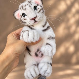 Puede incluir: Un juguete de peluche de cachorro de tigre blanco con rayas negras, ojos azules y largos bigotes blancos. El animal de peluche presenta pelaje blanco suave, orejas con tonos rosados y una pequeña nariz rosa, con cara detallada.