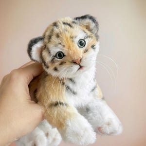 Puede incluir: Un peluche de cachorro de tigre de aspecto realista. El cachorro tiene un cuerpo crema y beige con rayas negras, ojos azules grandes y patas blancas. El juguete está diseñado para parecerse a un cachorro de tigre real.