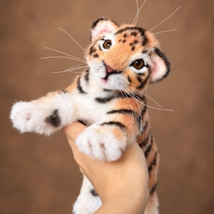 Può includere: Un cucciolo di tigre di peluche con pelliccia arancione, nera e bianca, caratterizzato da occhi marroni realistici, un piccolo naso rosa e lunghi baffi bianchi. Il peluche ha un aspetto morbido e strutturato con strisce nere dettagliate.