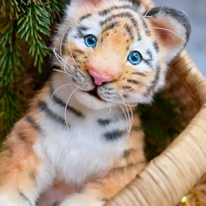 Può includere: Un cucciolo di tigre di peluche con strisce arancioni e nere, occhi blu e naso rosa, adagiato in un cestino intrecciato. Il cucciolo è circondato da aghi di pino verdi, creando un ambiente naturale. Il cestino è in materiale intrecciato marrone chiaro.