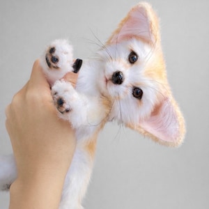 May include: A plush toy fennec fox with large ears and a white and tan coat. The fox is being held up by a hand, showcasing its small black paws and detailed facial features, including dark eyes and whiskers. The toy has a soft, fluffy appearance.