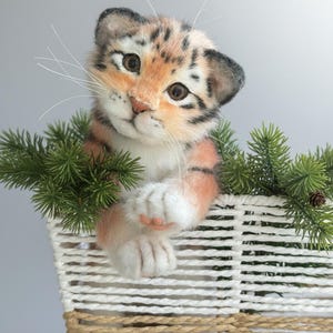 Pode incluir: Um filhote de tigre de pelúcia com listras laranja e pretas, aninhado em uma cesta branca tecida adornada com galhos de pinheiro verdes. O filhote tem uma expressão doce e é um item decorativo charmoso.