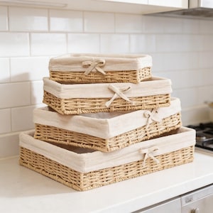 Peut inclure: Quatre paniers rectangulaires tressés de différentes tailles sont empilés. Chaque panier a une doublure en tissu clair et un nœud décoratif. Les paniers sont faits d'un matériau naturel brun clair.