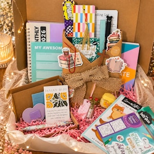 May include: A gift box filled with stationery items, including notebooks, sticky notes, bookmarks, and pens. The box contains a variety of colors, including pink, purple, and green. The box is decorated with a burlap bow and pink paper filler.