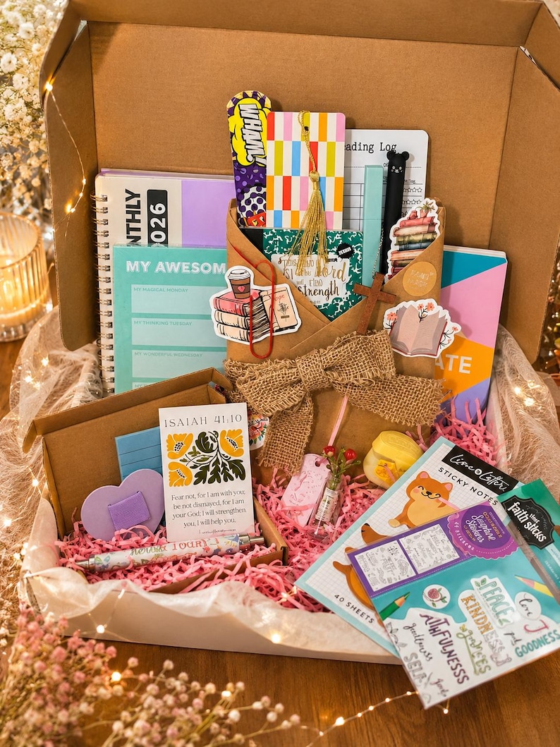 May include: A gift box filled with stationery items, including notebooks, sticky notes, bookmarks, and pens. The box contains a variety of colors, including pink, purple, and green. The box is decorated with a burlap bow and pink paper filler.