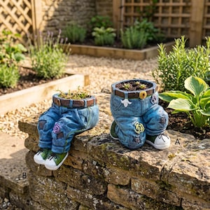 May include: Two blue denim jean-shaped planters with brown belts and white sneakers. One planter is in a seated position with legs extended, while the other is in a kneeling position. Both feature small floral accents and green plants inside.