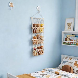 May include: A macrame wall organizer with three shelves, filled with plush teddy bears in various colors. The organizer hangs on a light blue wall, creating a cozy nursery or playroom decor.