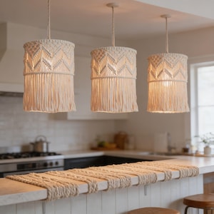 May include: Three off-white macrame pendant lights hang above a kitchen counter. Each light has a cylindrical shade with a geometric pattern and fringe. The lights are suspended from the ceiling by matching cords, casting a warm glow.