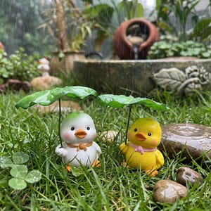 May include: Two small duck figurines standing in green grass during a rain shower. One duck is white with a light green head and orange scarf, while the other is yellow with a pink scarf. Each holds a green leaf overhead as a makeshift umbrella.