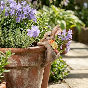Puede incluir: Una figura de conejo marrón con una mochila amarilla y roja colgada del borde de una maceta de terracota grande y desgastada, llena de vibrantes flores moradas en forma de campana y follaje verde en un jardín.